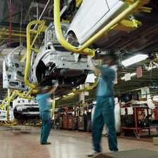 Two workers wearing green shirts and blue pants work underneath a car in an automobile factory. There are assembly lines, partially constructed cars, tools and electrical equipment in the background.