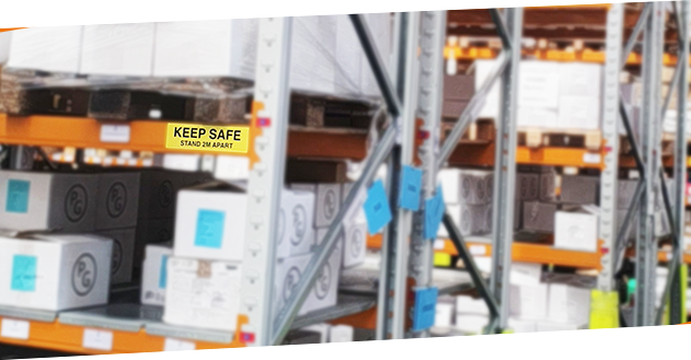 An image of PG-labelled boxes on warehouse racking. A safety label on the racking, which reads "KEEP SAFE, STAND 2M APART" is in focus.
